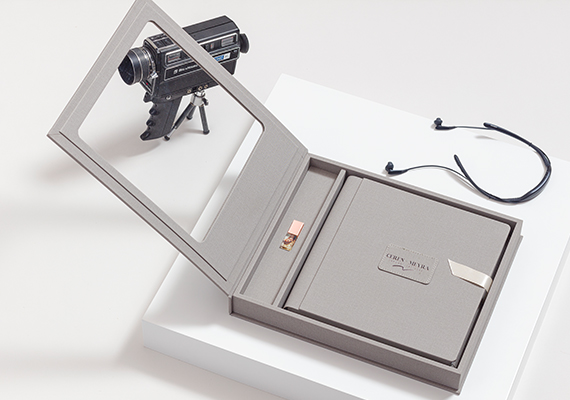 person writing in a notebook beside by an iPad, laptop, printed photos, spectacles, and a cup of coffee on a saucer