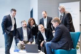 man wearing a black jacket, white shirt, blue jeans, and brown boots, playing a white electric guitar while sitting on an amp